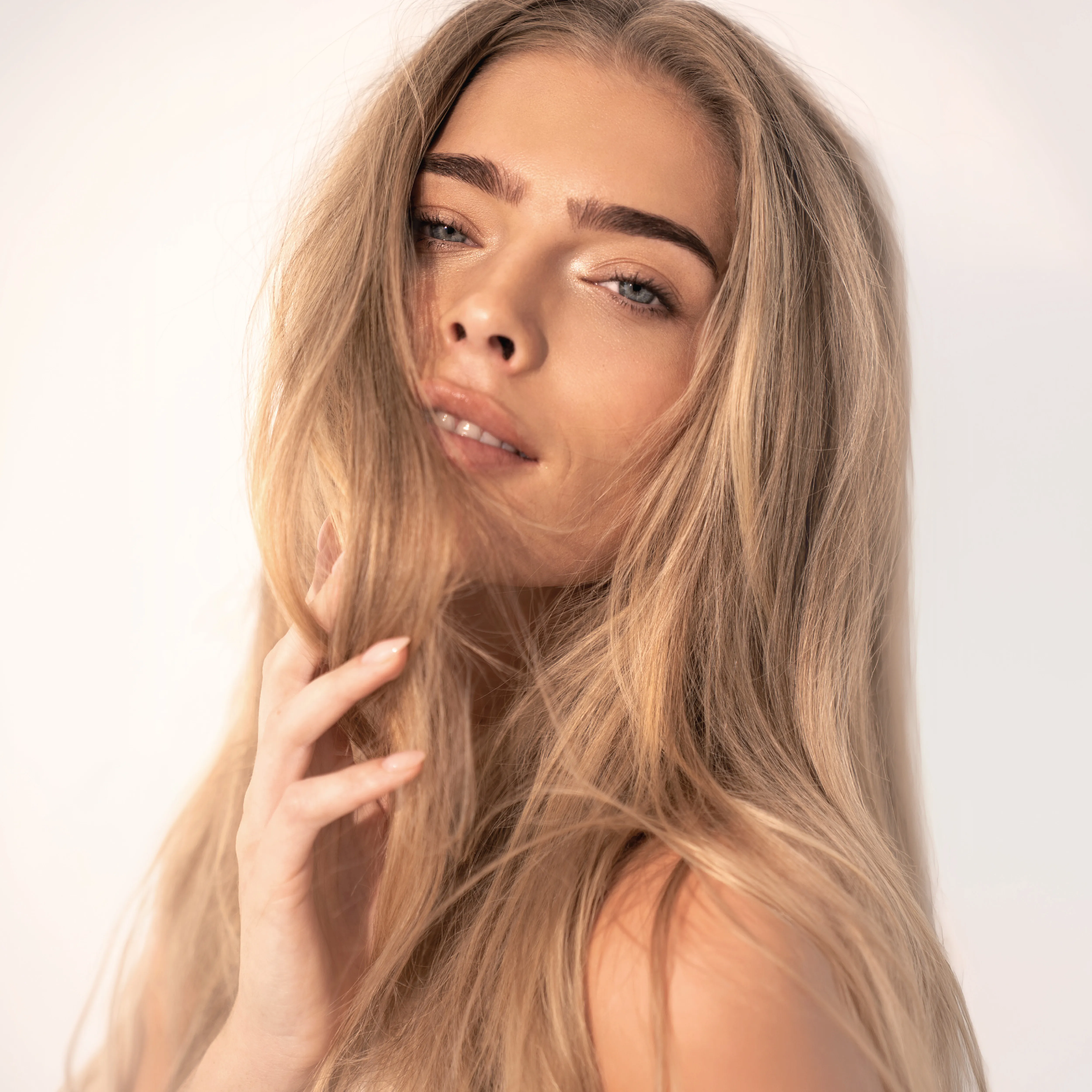 Frau mit langen, gl&auml;nzenden blonden Haaren und nat&uuml;rlichem Make-up vor hellem Hintergrund.